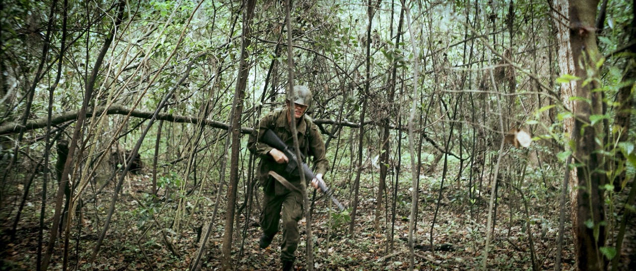 a man in a war uniform runs through a dense forrest