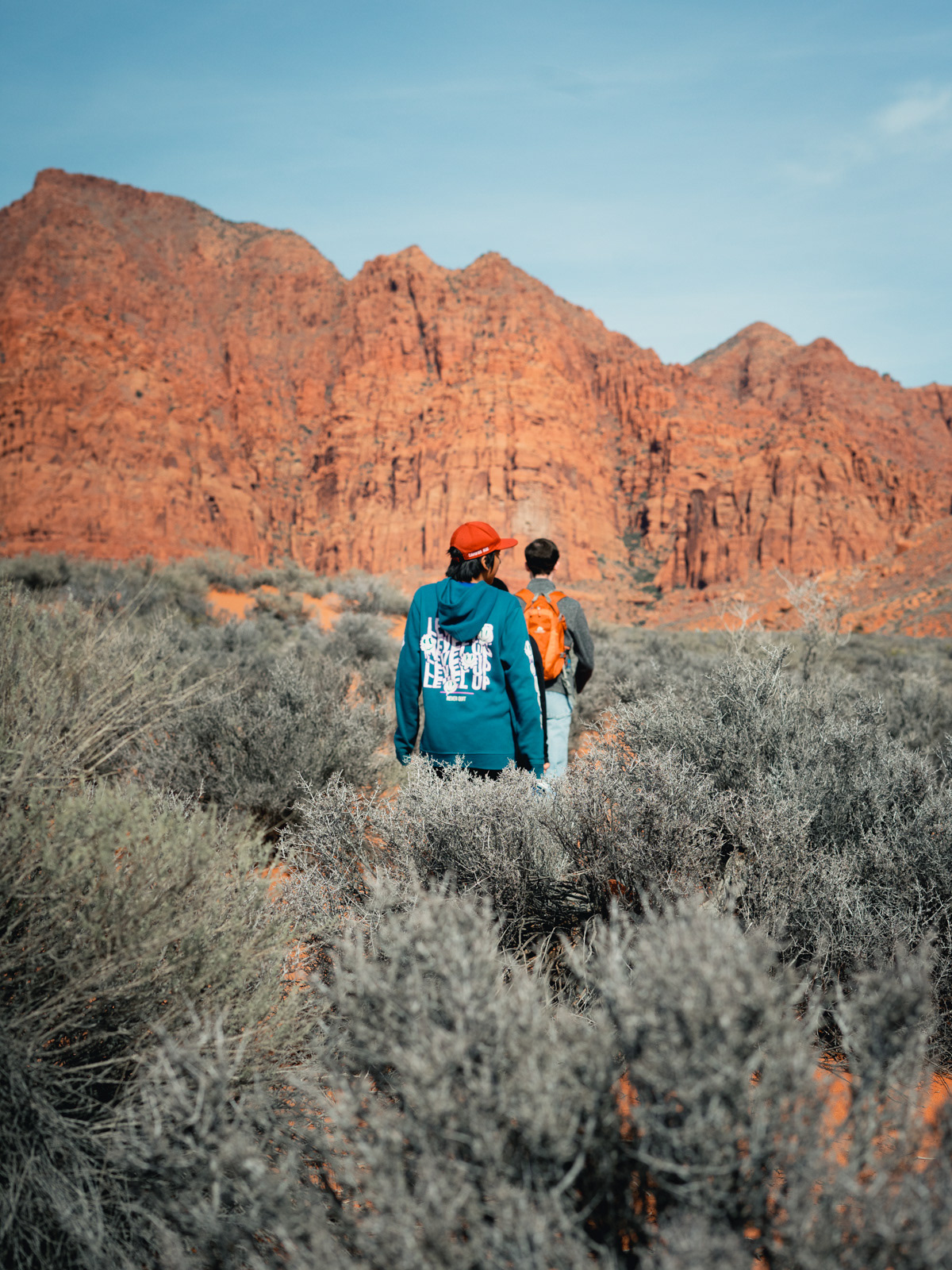 two people walk into a brush field towards a red rock wall